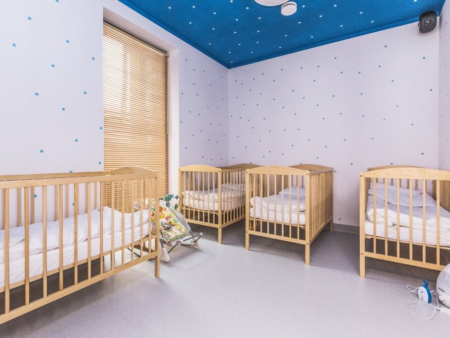 Small hotel nursery with five wooden cribs, star-patterned blue ceiling, soft white bedding, window blinds and a baby bouncer.