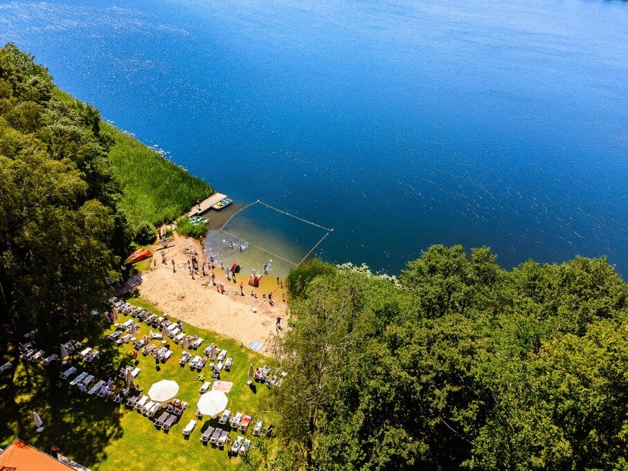 Lakeside beach with roped swimming area, docked kayaks, sun loungers and umbrellas on a grassy lawn by forest.