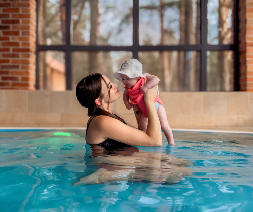 Kobieta trzymająca niemowlę nad wodą na hotelowym basenie, duże okna z widokiem na las i ceglana atmosfera spa