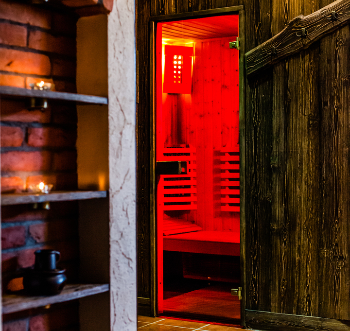Wood-paneled sauna with warm red lighting visible through an open door, rustic wooden walls, tiled floor and candle-lit brick shelving.