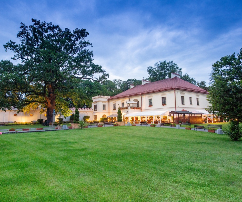 Zabytkowy dwór z oświetlonym tarasem restauracyjnym, zadbanym trawnikiem, ławkami i potężnym dębem o zmierzchu