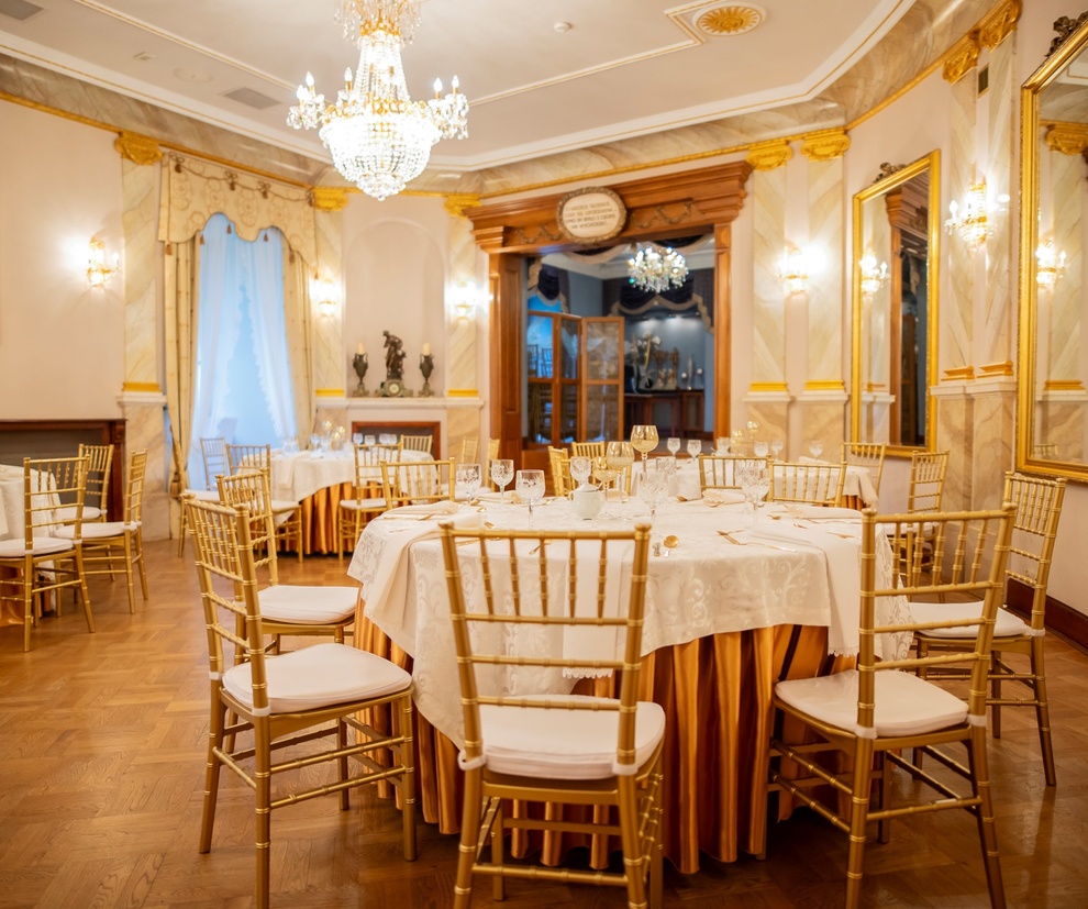 Elegancka sala bankietowa z kryształowym żyrandolem, złotymi krzesłami chiavari i okrągłymi stołami nakrytymi białymi obrusami
