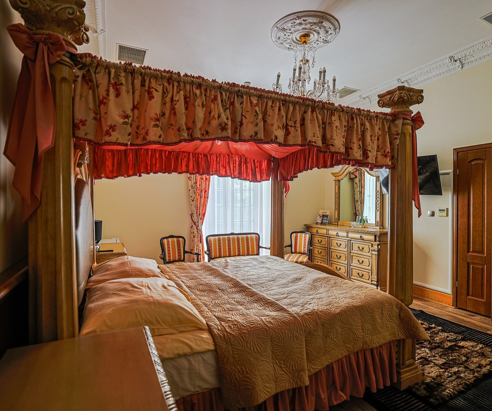 Baldachimowe łóżko z kwiatowymi zasłonami, kryształowy żyrandol, rzeźbiona komoda i pasiaste fotele w eleganckim apartamencie.