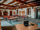Historyczna sala konferencyjna w hotelu z drewnianymi belkami, żyrandolami, ceglaną ścianą i kominkiem; stoły w rzędach, czerwone krzesła, laptop i woda.