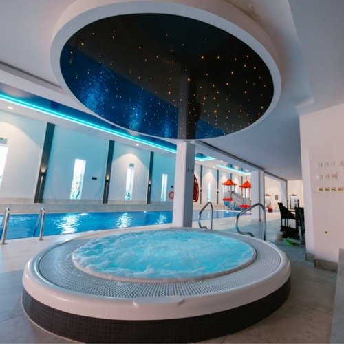 Innen-Spa mit sprudelndem Rund-Whirlpool, langem Schwimmbecken, Sternenhimmel an der Decke, blauer LED-Beleuchtung und Natursteinwand.