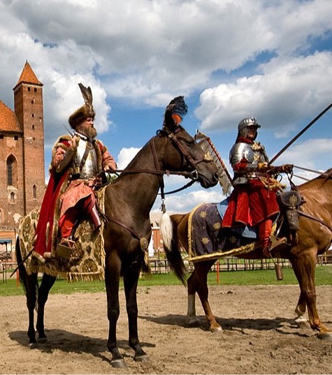 Two mounted knights in period armor perform in a castle courtyard before a large red-brick medieval castle, spectators seated.
