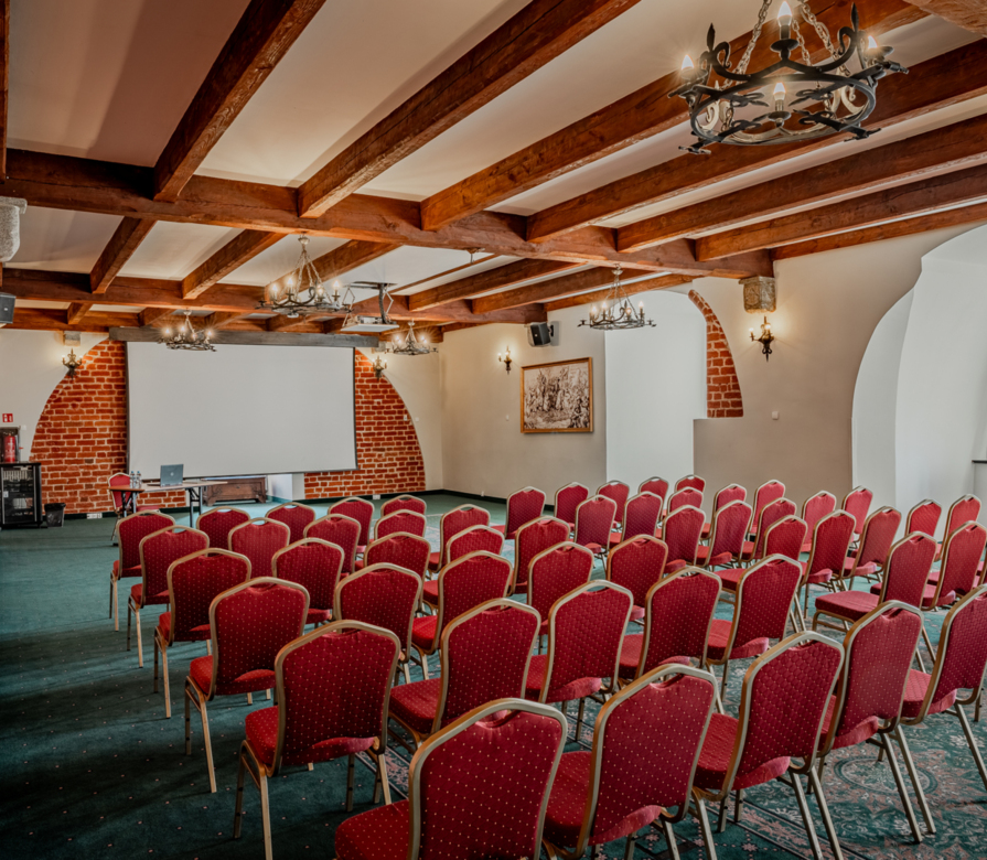 Sala konferencyjna w historycznym hotelu z odsłoniętymi ceglanymi łukami, drewnianymi belkami, żyrandolami, czerwonymi krzesłami i ekranem projekcyjnym