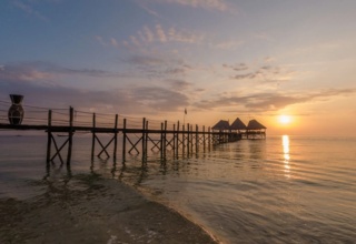 Wooden pier extending over calm ocean waters toward thatched-roof huts at sunset, highlighting peaceful beachfront ambiance