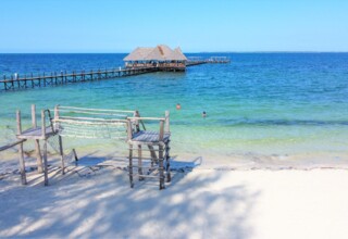 Secluded sandy beach with wooden playground, clear turquoise water, and a pier leading to thatched-roof overwater restaurant