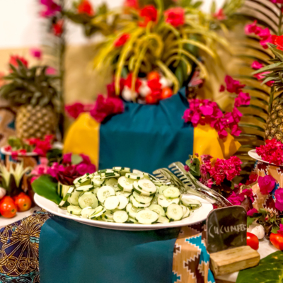 Fresh cucumber slices on a white plate surrounded by tropical decor with pineapples, tomatoes, and vibrant pink flowers