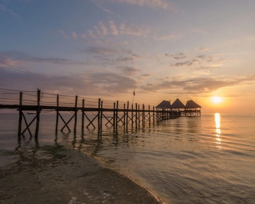 Wooden pier extending over calm ocean waters toward thatched-roof huts at sunset, highlighting peaceful beachfront ambiance
