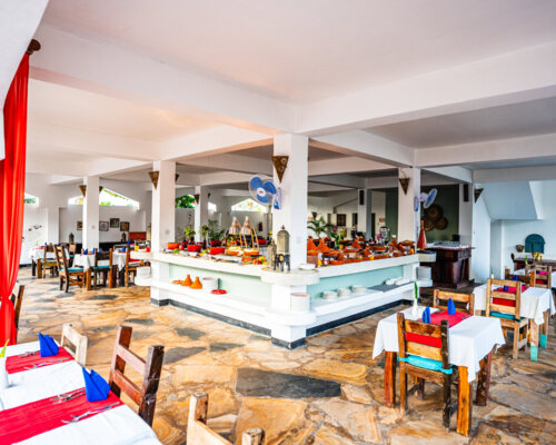 Bright hotel dining area with rustic wooden chairs, white tablecloths, red runners, blue napkins, and a central buffet counter