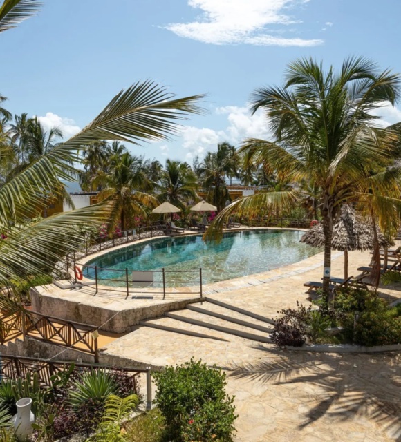 Tropical resort pool surrounded by palm trees, sun loungers, umbrellas, and a stone-paved walkway leading to white balcony rooms