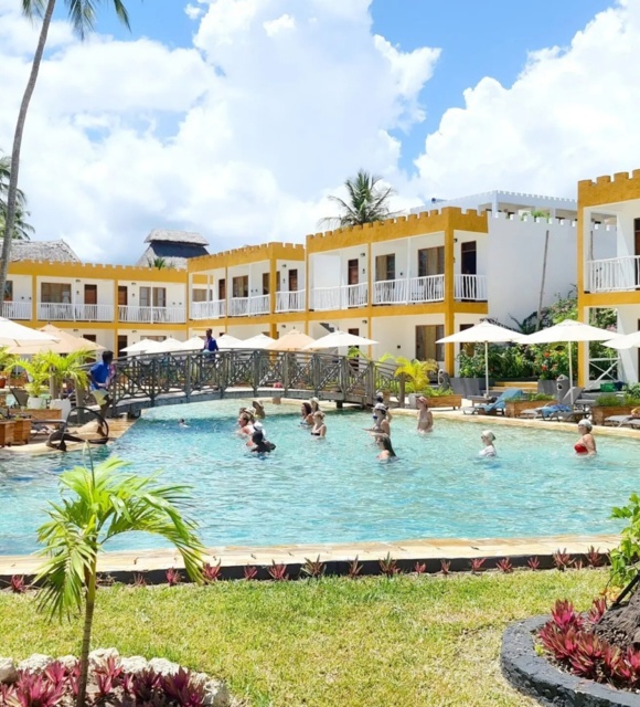 Resort pool with guests enjoying water activities, surrounded by two-story yellow and white rooms, palm trees, and sun umbrellas