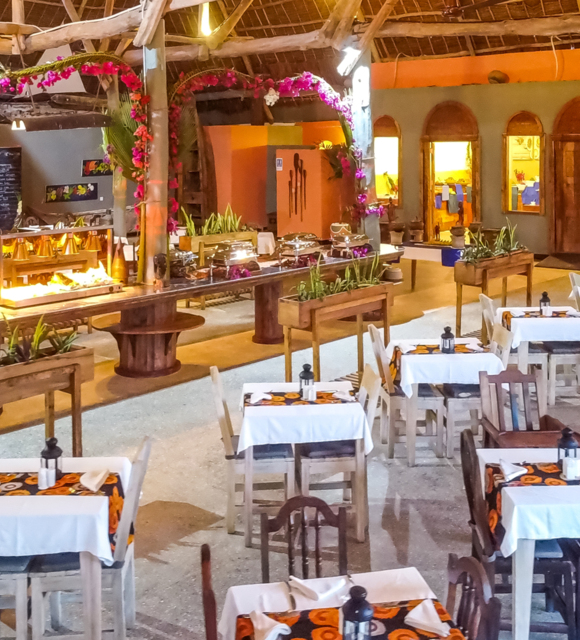 Rustic open-air restaurant with wooden tables covered in white linens, decorated buffet with flowers, and warm ambient lighting