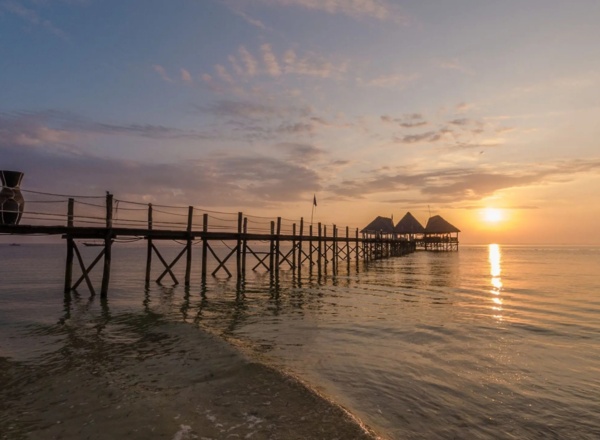 Wooden pier extending over calm ocean waters toward thatched-roof huts at sunset, highlighting peaceful beachfront ambiance