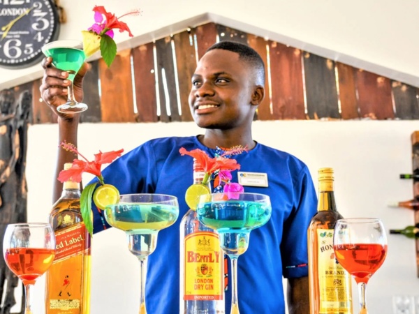 Smiling bartender in blue uniform holds tropical cocktail garnished with flowers and pineapple at stylish hotel bar