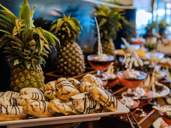 Tray of cream-filled eclairs with white and dark chocolate drizzle beside fresh pineapples and layered dessert glasses in buffet setting