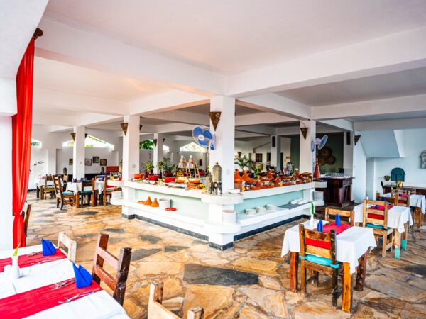 Bright hotel dining area with rustic wooden chairs, white tablecloths, red runners, blue napkins, and a central buffet counter