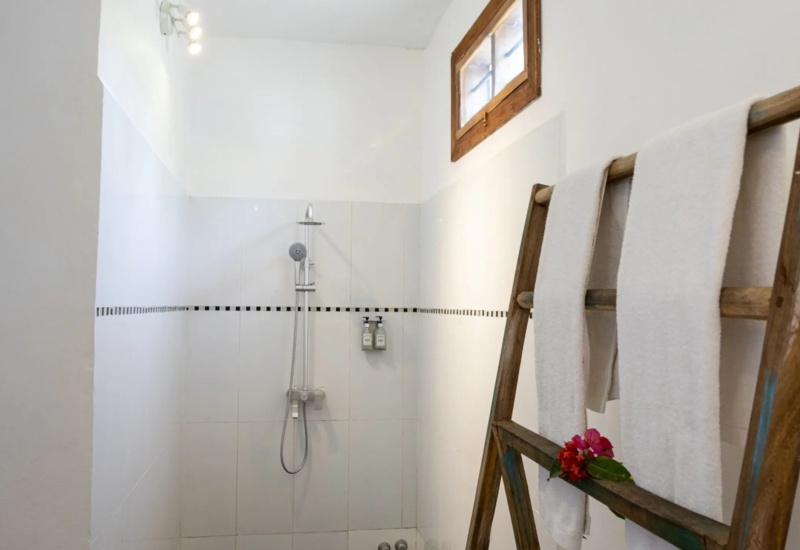 Minimalist white-tiled shower area with rainfall showerhead, wooden towel ladder with white towels and fresh flowers, small window