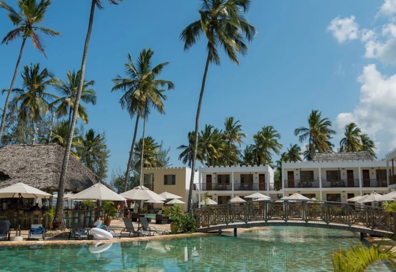 Tropical resort pool with sun loungers, umbrellas, a thatched poolside bar, palm trees, and white two-story guest rooms with balconies