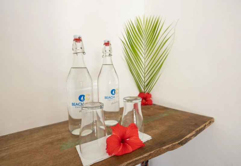 Two glass bottles of water and two upside-down glasses on a wooden shelf, decorated with red hibiscus flowers and a green palm leaf