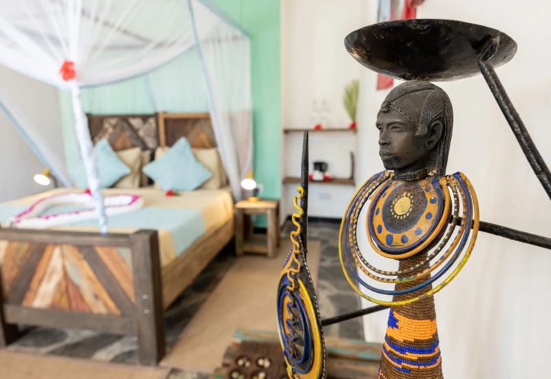 Rustic hotel room with wooden bed, soft blue pillows, mosquito net, and colorful handcrafted African sculpture in foreground