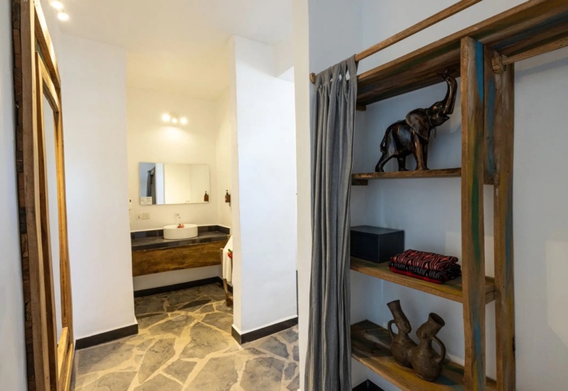 Bathroom area with stone flooring, wooden-framed mirror, open shelf with decorative elephant statue and pottery, and modern sink.