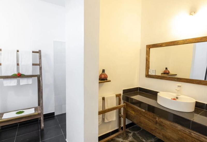 Bathroom with modern black tiled counter, white oval sink, large wooden-framed mirror, rustic towel rack holding white towels with decorative flowers.