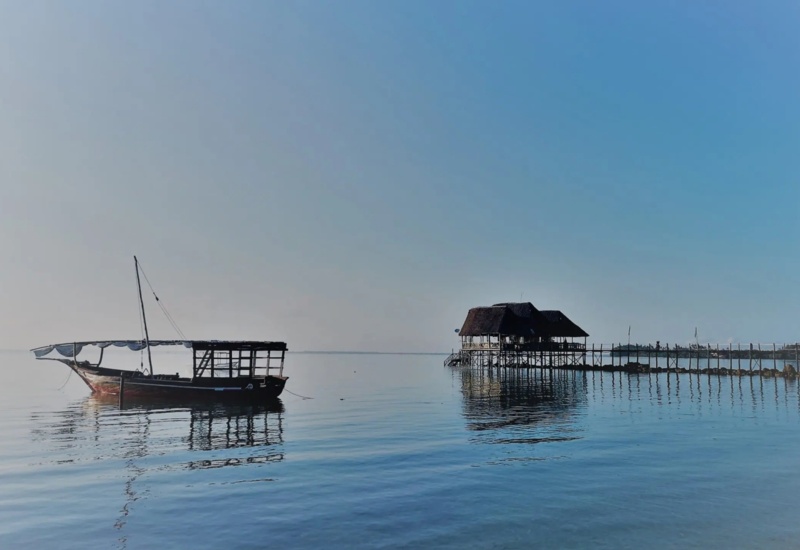 Traditional wooden boat floating on calm water near a thatched-roof pier house under clear blue sky
