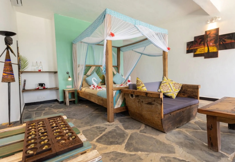 Cozy room with wooden four-poster bed draped in white and blue fabric, rustic sofa with patterned pillows, stone floor, and artistic decor.