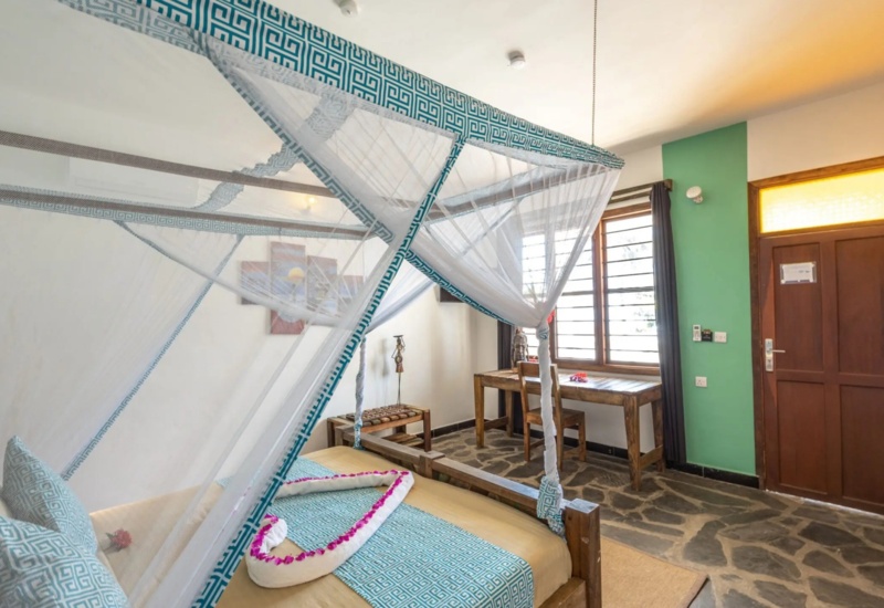 Cozy hotel room with a canopy bed draped in mosquito netting, patterned blue accents, stone floor, and wooden desk by window.