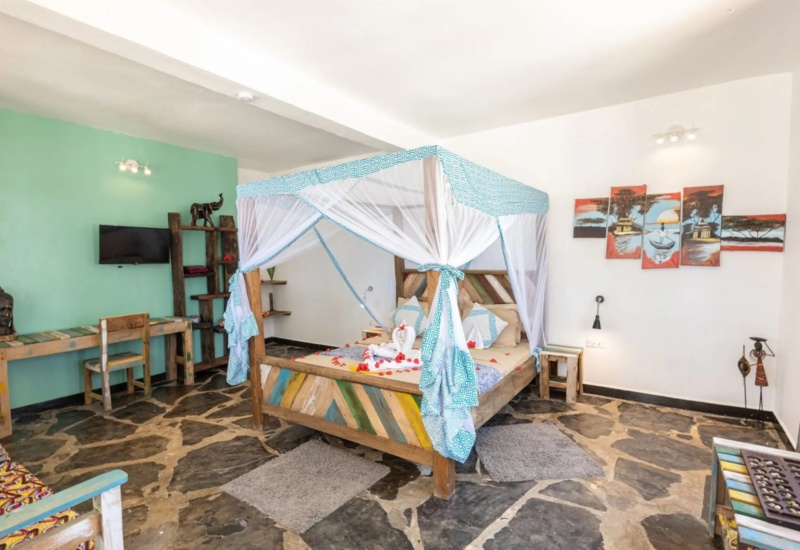 Bright room with a colorful four-poster bed draped in white and blue mosquito netting, rustic furniture, and stone flooring.