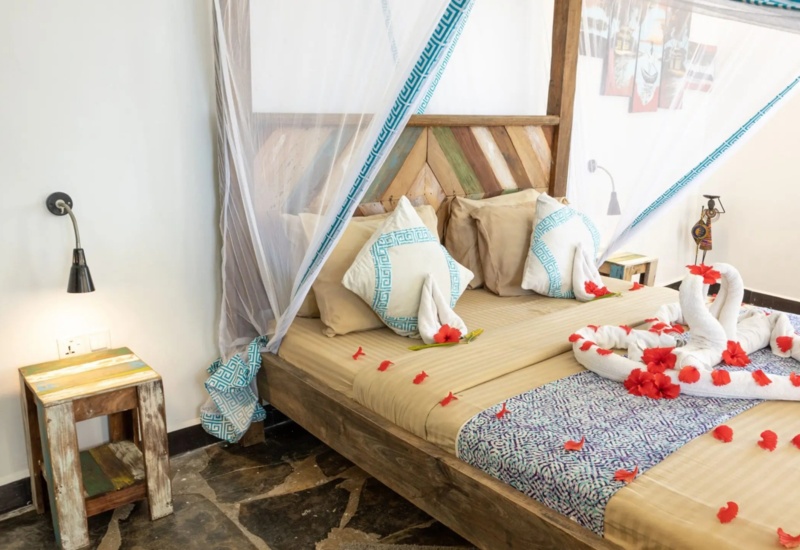 Rustic wooden bed with beige linens, decorative pillows, mosquito netting, and towel swans adorned with red flower petals.