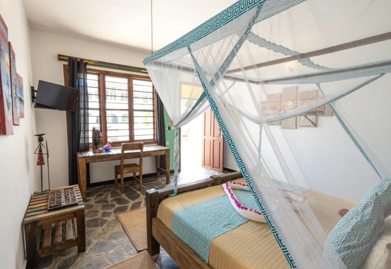 Cozy hotel room with wooden bed draped in mosquito net, patterned teal bedding, rustic desk, and stone flooring in natural light