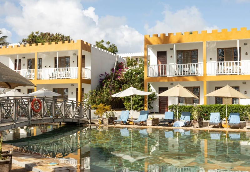 Two-story hotel with bright yellow trim, balconies overlooking a serene pool surrounded by sun loungers and umbrellas