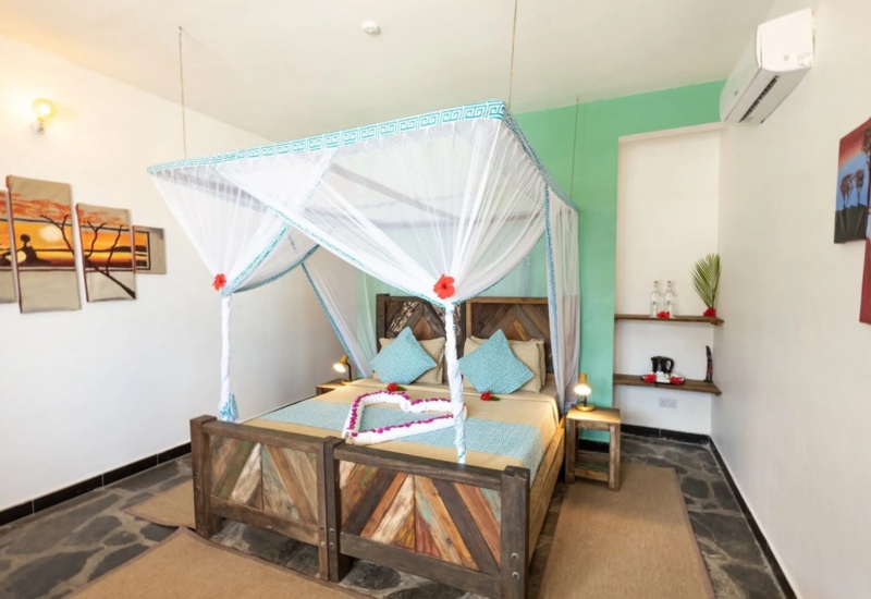 Cozy room with wooden canopy bed draped in white netting, turquoise accent wall, bedside lamps, and vibrant local artwork