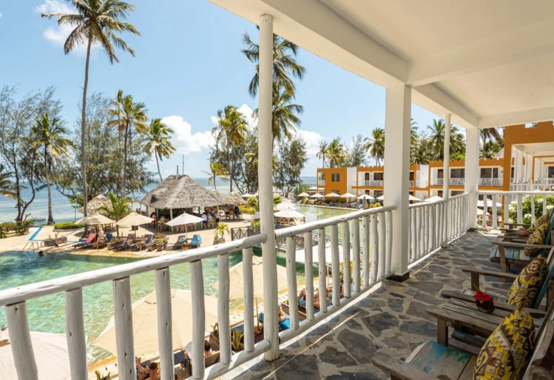 Oceanfront balcony with rustic wooden chairs overlooking pool, thatched pool bar, palm trees, and beachfront resort under clear sky
