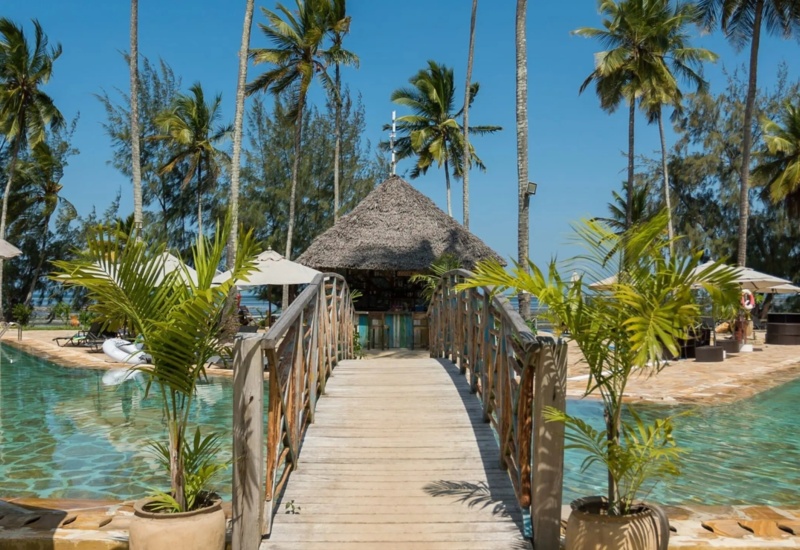 Wooden bridge crosses turquoise pool leading to a thatched-roof pool bar surrounded by palm trees and lounge chairs