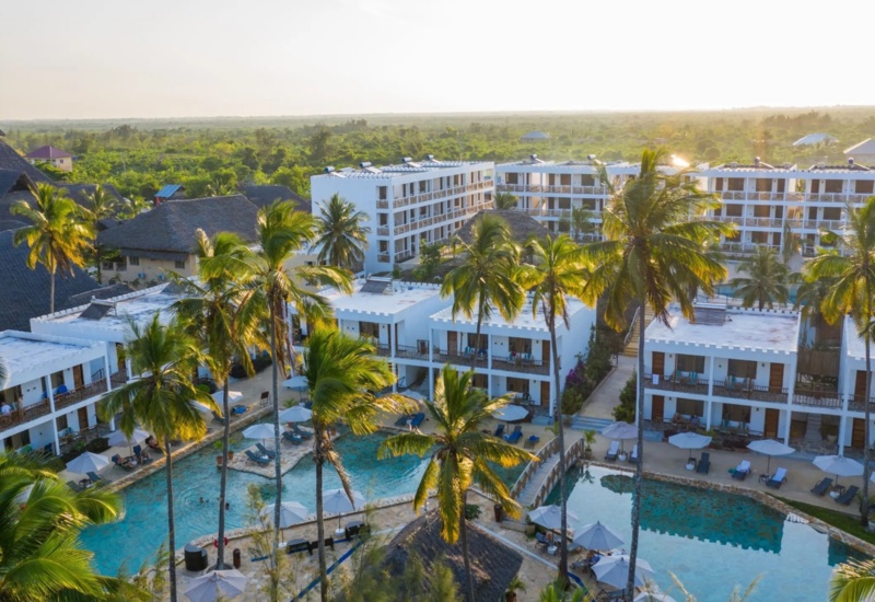 Tropical resort with white modern buildings, palm trees, and a large pool surrounded by sun loungers and umbrellas during sunset