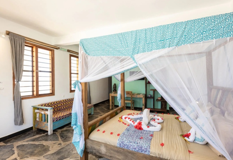 Bright room with a four-poster bed draped in white and blue mosquito netting, decorated with towel swans and rose petals, stone floor, and wooden furnishings.