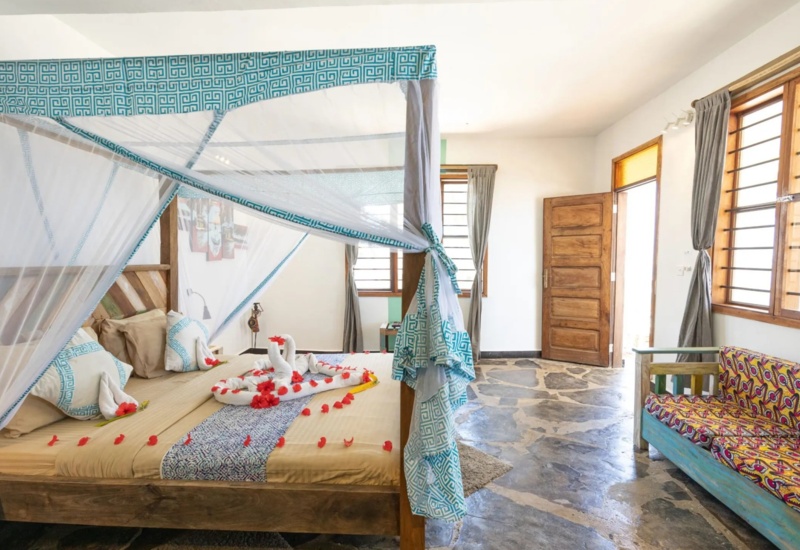 Bright room with wooden four-poster bed draped in patterned mosquito netting, flower petal decorations, and a colorful sofa on stone flooring.