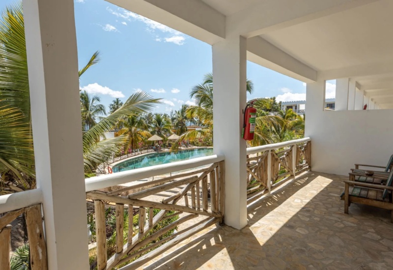 Sunlit balcony with rustic wooden railing, two wooden chairs, overlooking tropical palm trees and a curved pool under blue sky