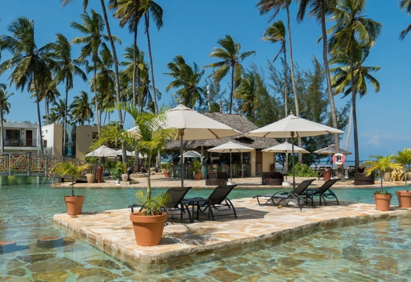 Tropical resort pool with lounge chairs and umbrellas on stone island, surrounded by palm trees and waterfront bar under a clear blue sky