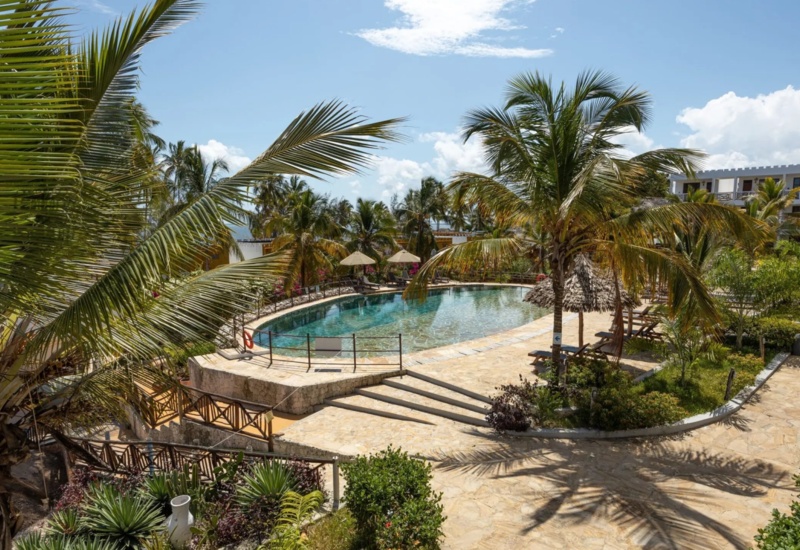 Tropical resort pool surrounded by palm trees, sun loungers, umbrellas, and a stone-paved walkway leading to white balcony rooms