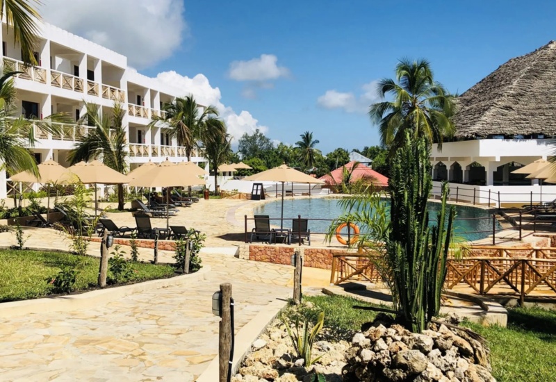 Resort pool area with modern white balconies, shaded loungers, palm trees, and a thatched-roof poolside bar under a sunny blue sky