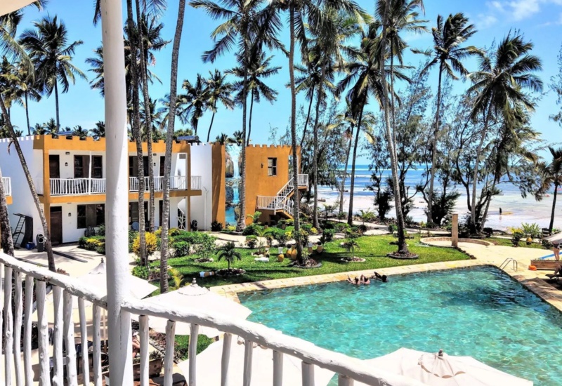 Tropical beachfront hotel with palm trees, two-story rooms with balconies, lush garden, and a clear blue swimming pool with guests relaxing