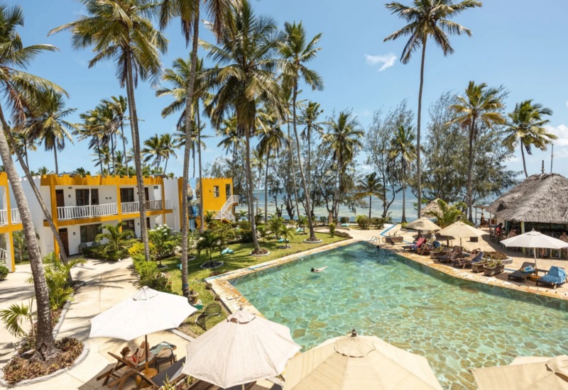 Tropical beachfront hotel with yellow-white two-story rooms, a large pool, palm trees, sun loungers, and a thatched-roof pool bar.