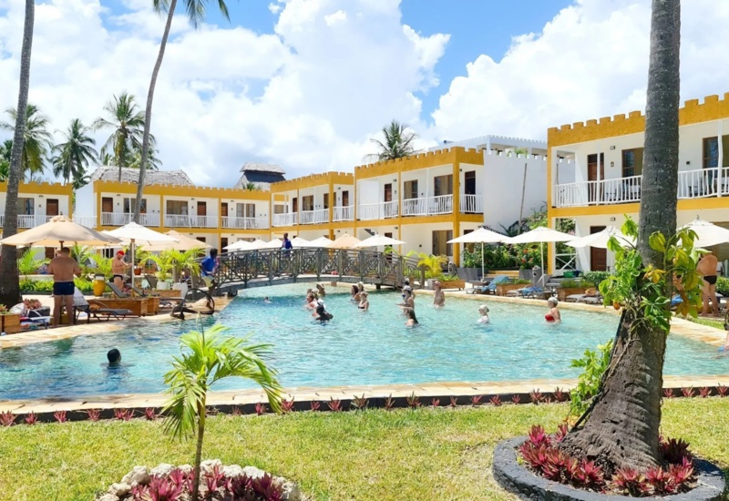 Resort pool with guests enjoying water activities, surrounded by two-story yellow and white rooms, palm trees, and sun umbrellas