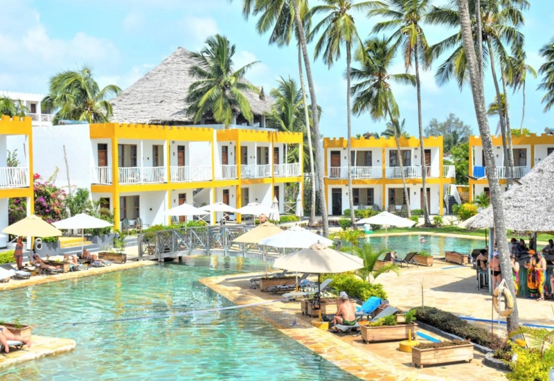 Resort pool surrounded by yellow-bordered white rooms, tall palm trees, sun loungers, umbrellas, and guests enjoying the poolside bar